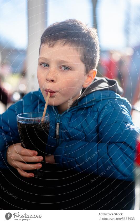 A young boy in a blue jacket looking at camera while sipping a dark soda through a straw at a table drink casual leisure refreshment beverage fizzy cola glass