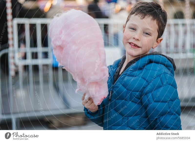 Cheerful young boy in a blue jacket looking at camera while holding huge cotton candy at an amusement park treat sweet fluffy outdoor child sugary dessert snack