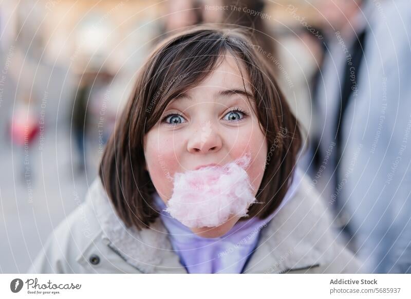 Close-up of excited girl with wide eyes looking at camera while enjoying a mouthful of cotton candy on a city blurred street background close-up sweet treat