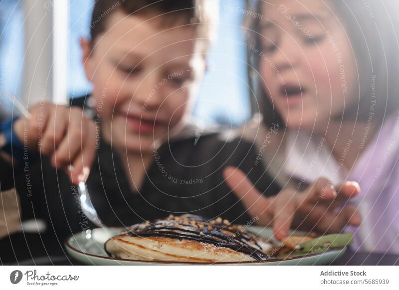 Two children enjoying a meal together, with a boy holding a fork over a pancake topped with syrup and a girl pointing towards it topping plate food sweet