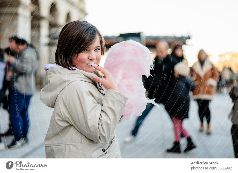 Side view of smiling girl in a beige coat playfully eating cotton candy on a busy street enjoying smile blurred background sweet treat dessert fluffy pink