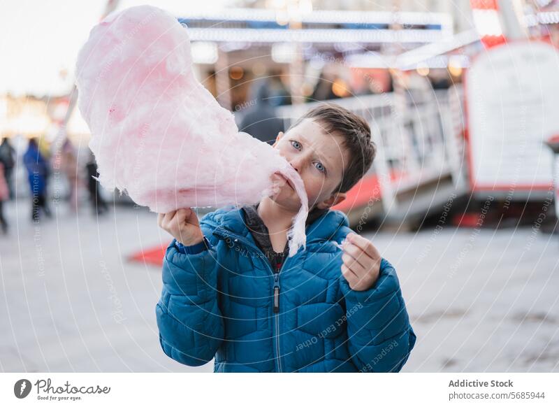 Boy in blue coat playfully eating cotton candy at a carnival with rides in the background looking at camera boy sweet treat dessert fluffy pink outdoor sugary