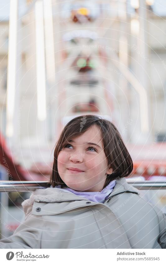 A girl in a beige coat with a dreamy expression looks up while sitting in front of a Ferris wheel amusement park child contemplative happy casual outdoor