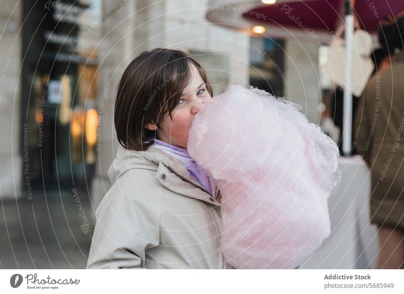 A girl with a beige jacket looking at camera while eating a large pink cotton candy on a city street outdoor sweet snack confection treat indulgence dessert