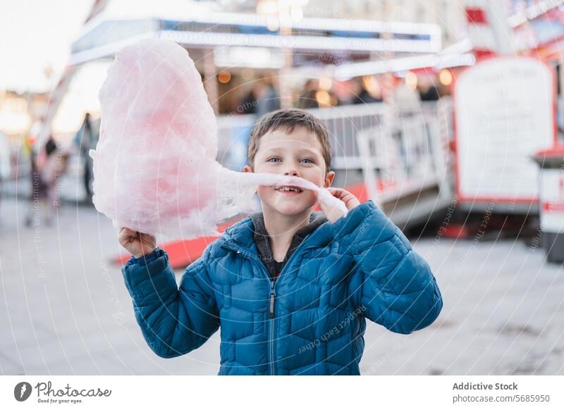 Happy delighted boy in a blue coat looking at camera while posing with a big piece of cotton candy at a fairground amusement sweet treat fluffy pink outdoor