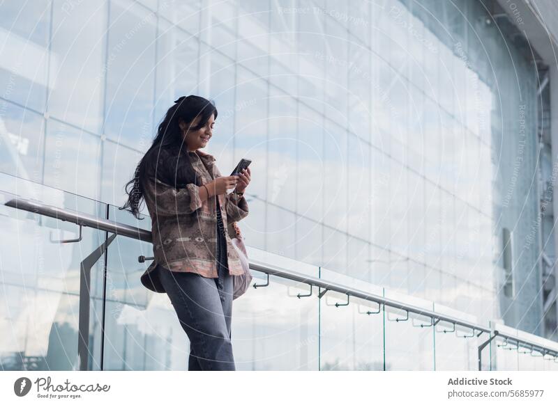 Smiling woman using smartphone in city cheerful smile handrail trousers modern glass wall young contemporary female surfing lean urban happy street gadget