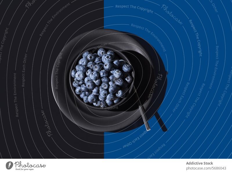 From above of bowl full of ripe blueberries accompanied by a spoon on a dual-tone blue and black background Blueberries fruit fresh healthy food dessert snack