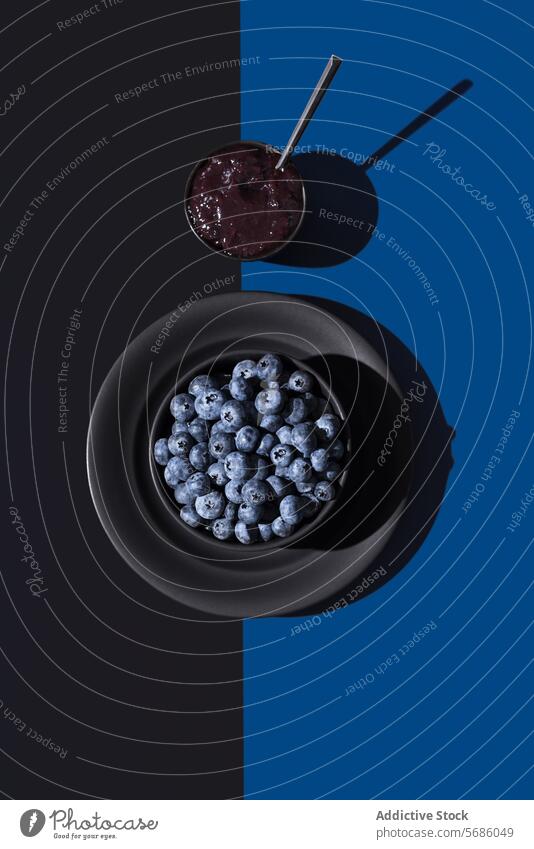 Fresh blueberries in a black bowl with a cup of blueberry jam on a split blue and black background, showcasing a play of light and shadow Blueberry spoon fresh