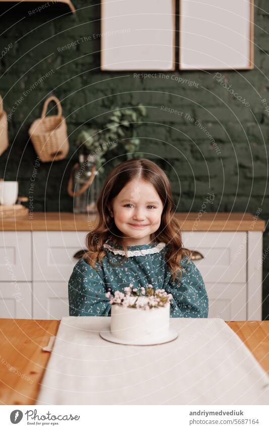 Adorable little child girl wears festive dress takes a bite out of a decorated flowers name cake at a birthday party. Happy smiling kid licks white cream from her dirty face and shows thumb up indoors
