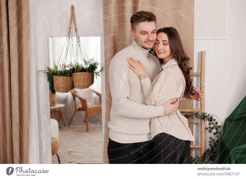 Happy young couple in love is hugging gentle and smiling together celebrating Valentine's day at cozy Scandinavian style home. Man and woman enjoying spending time together, relaxing on date.
