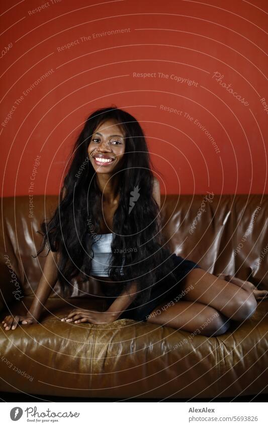 Young, slim woman with long black hair and hot pants sits crosswise on a brown couch in front of a red wall and smiles into the camera Woman Young woman