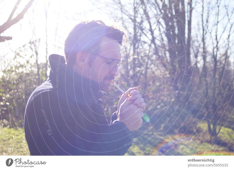 Man lights a cigarette outdoors and is illuminated by the winter sun smoking Smoking Back-light Glare Cigarette Ignite Exterior shot Meadow shrubby hands stop