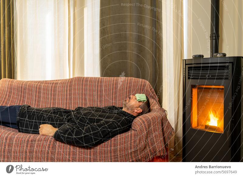 Sick Hispanic man lying on the couch with a robe and a cloth on his forehead in the heat of a pellet stove. sick male sofa fever adult illness health sickness