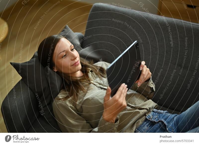 Relaxed young woman watching video on digital tablet, lying on a couch in the living room technology winter using home online shopping communication one person