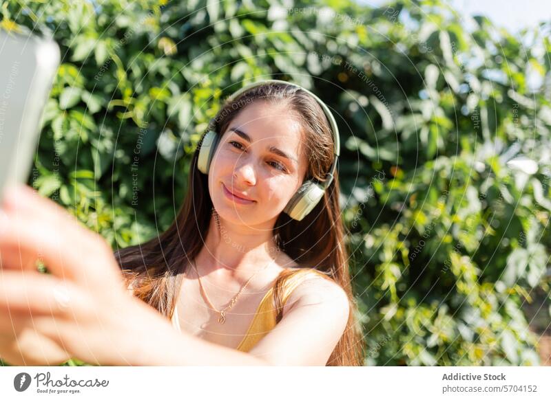 Young woman taking selfie with smartphone outdoors young music headphones garden sunny technology mobile photo smiling leisure happiness recreation enjoyment