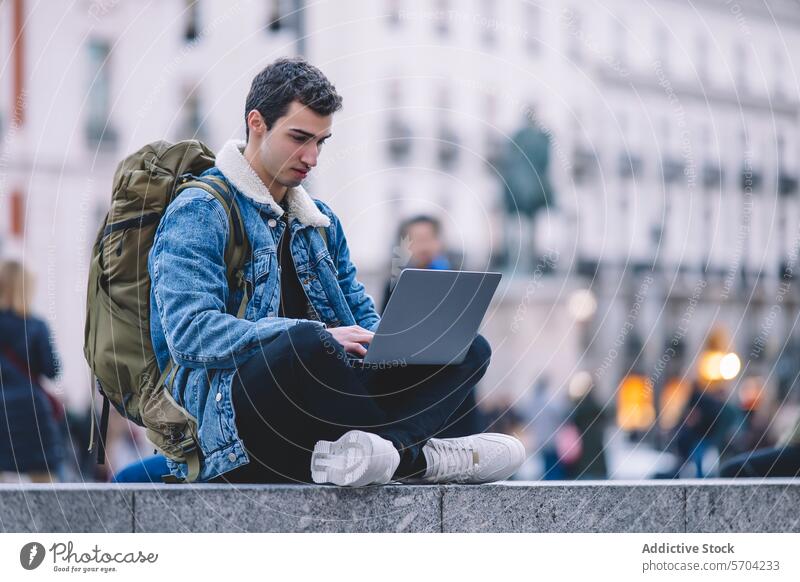 Young man works on his laptop while seated on the steps in a lively Madrid square young working outdoor city life backpack denim jacket urban male adult