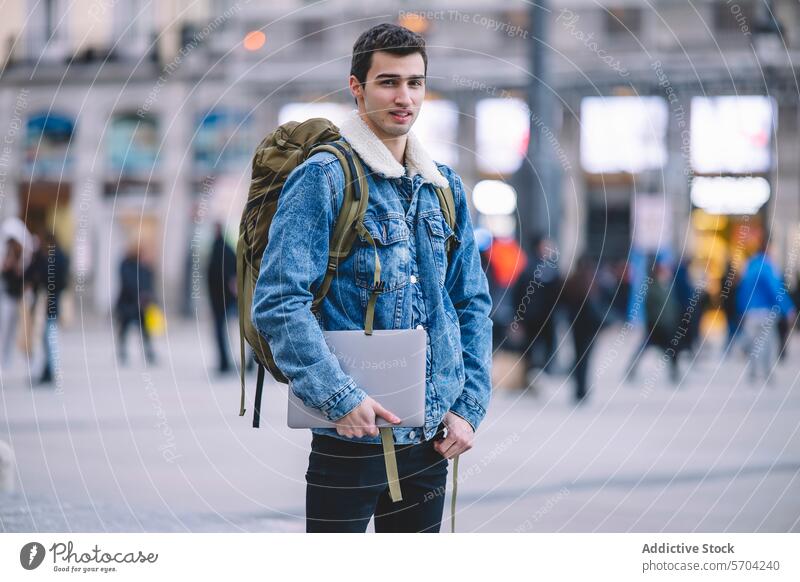 Confident young man holding a laptop stands in a bustling Madrid square professional confident backpack denim jacket urban cityscape male adult technology