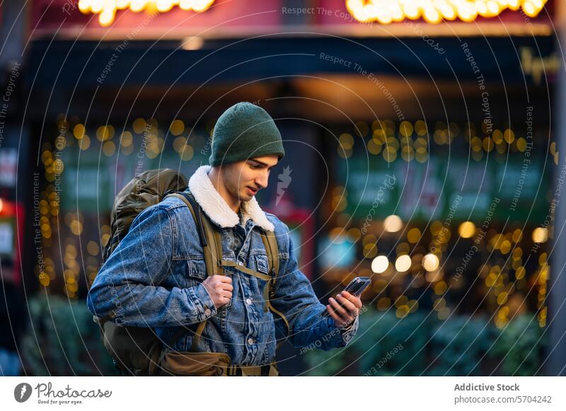 Young man in a beanie and denim jacket using his phone, with twinkling city lights in the background young bokeh urban evening mobile browsing texting casual
