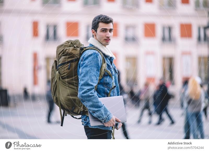 Side view of young man with backpack and laptop looking back on a busy Madrid street tech-savvy traveler denim jacket urban city male adult technology portable