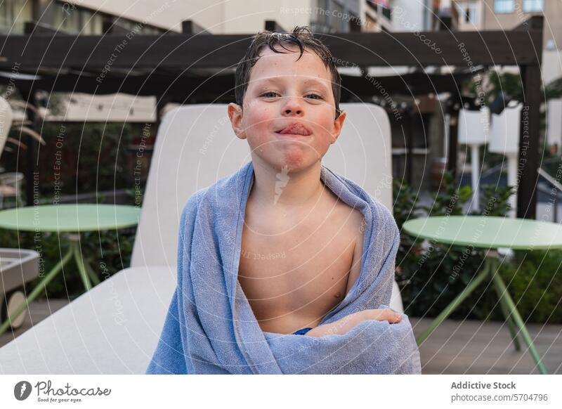 Young boy wrapped in towel after swimming, smirking child pool outdoor leisure summer daytime kid blue enjoyment fun happy cheerful wet hair sun lounging patio