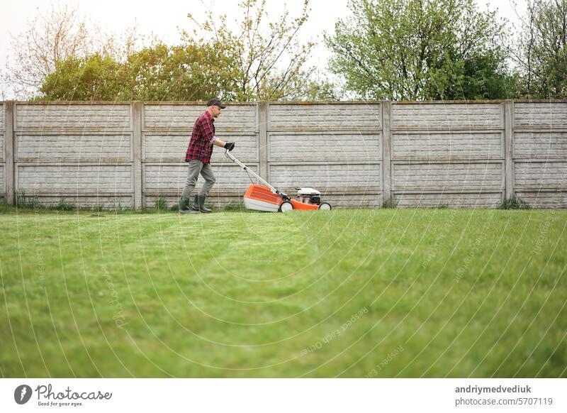 view of man in casual clothes mows lawn with lawn mower at backyard of his house. Husband takes care of garden on spring cloudy day. Modern gasoline garden equipment. Landscaping work