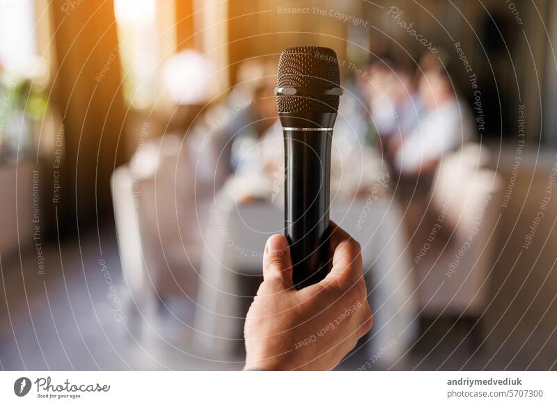 Unrecognisable male hand holds black microphone, master of ceremony or showman speech to audience in event. On background sit people guests by the table. Party and festive events concept.