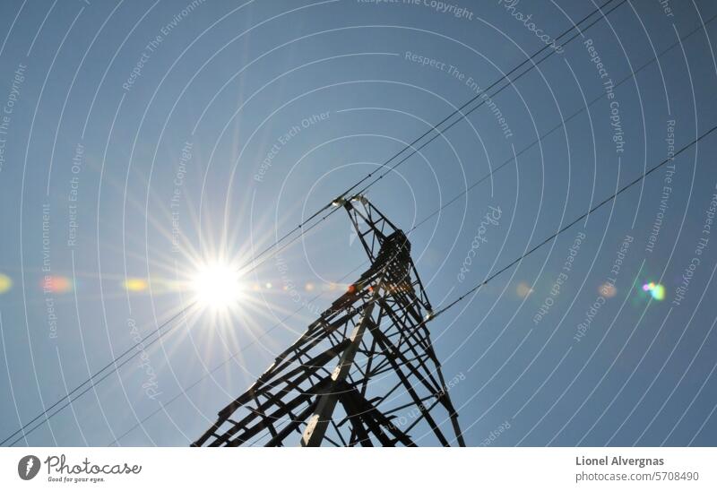 Very graphic composition with the sun along electrical wires on the top of a big electrical pylon with a blue sky electricity cable flare glare metal tall high