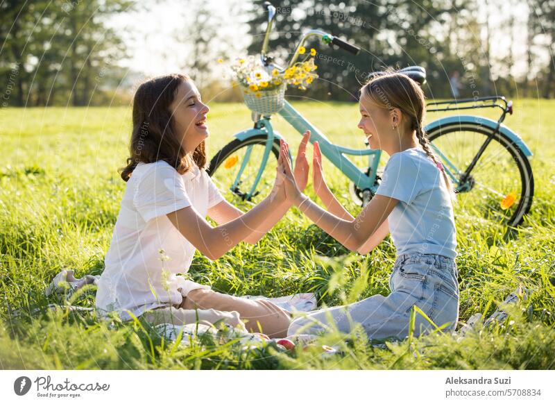 Two teenage girls spend time on green grass lawn in park, braid pigtails and tails for each other, enjoy summer and vacations attractive beauty bicycle bike