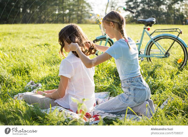 Two teenage girls spend time on green grass lawn in park, braid pigtails and tails for each other, enjoy summer and vacations attractive beauty bicycle bike