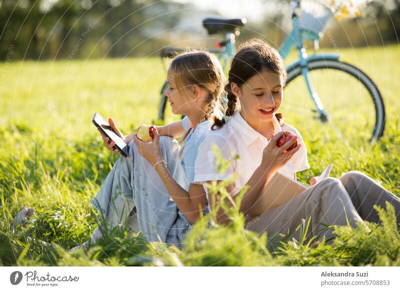 Two girls spend time on green grass lawn in park together, scroll through smartphones, read books, eat apples, enjoy summer and vacations beautiful bike casual