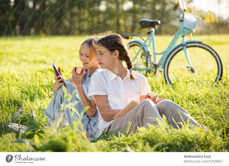 Two girls spend time on green grass lawn in park together, scroll through smartphones, read books, eat apples, enjoy summer and vacations beautiful bike casual