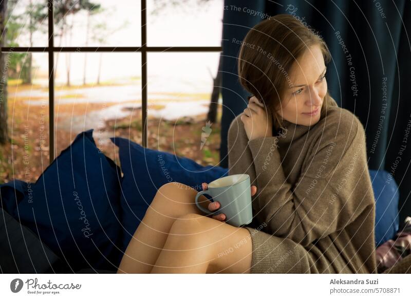 Pretty young woman reading book and drinking morning coffee at home sitting at the window behind which there is an autumn landscape adult background beautiful