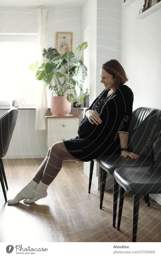 Lateral portrait of a young, pregnant, laughing woman sitting on a chair in the living room Woman Young woman Pregnant pregnancy pregnant woman Sit Laughter Joy