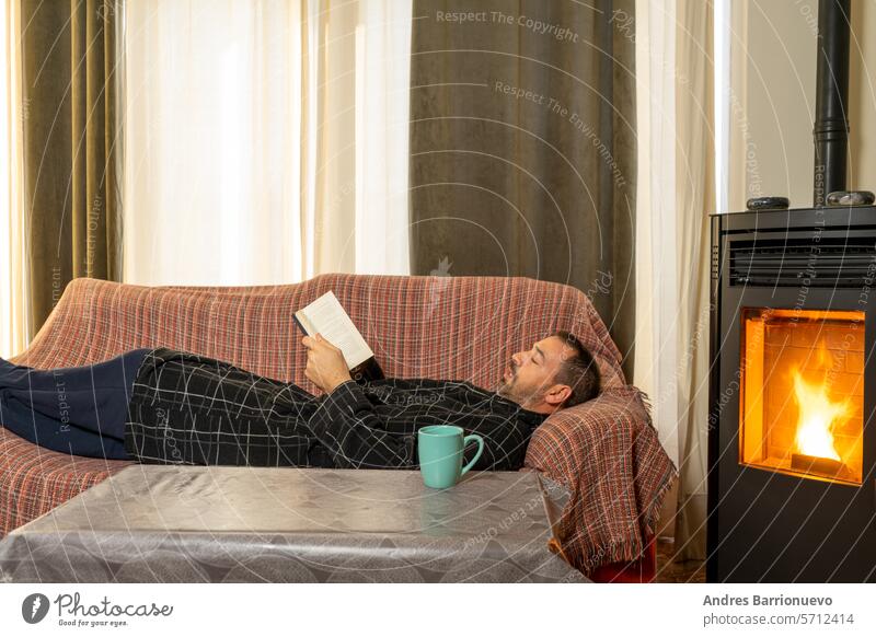 Hispanic man with a beard in his 40s lying on the couch reading a book, he is very comfortable with the heat of a pellet stove ifestyle adult interior leisure