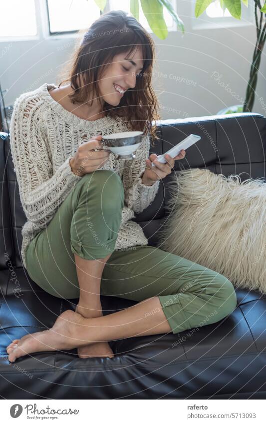 A joyful woman on a sofa with a bowl and a smartphone, embodying comfort and modern living. knit-sweater green-pants barefoot relaxation home cozy technology
