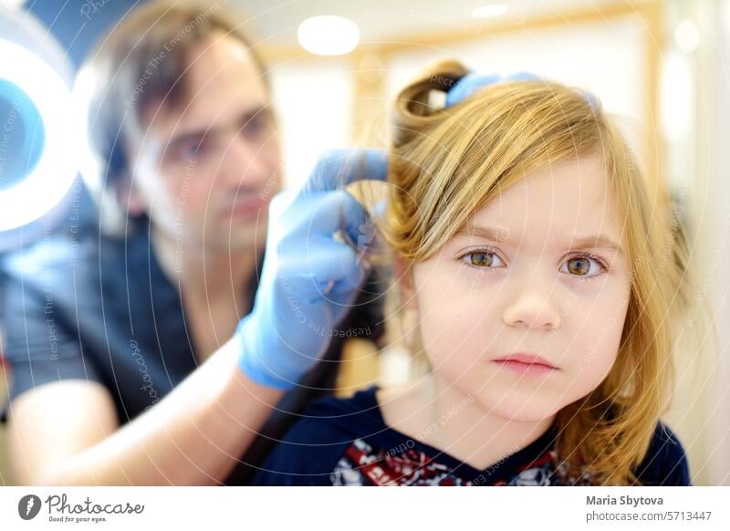 Close-up portrait of little girl during appointment of dermatologist in modern clinic. Doctor examines child hair and scalp for lice and nits. Pediculosis is common disease in kids groups