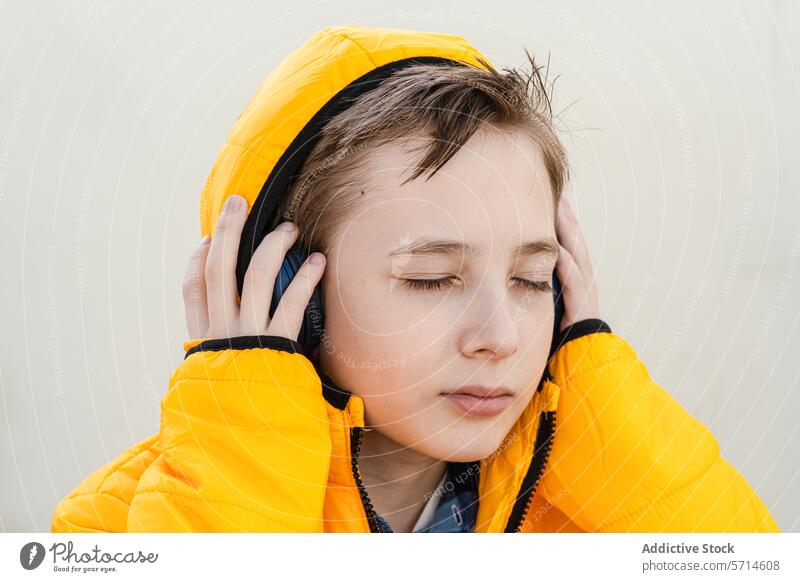A contemplative young boy in a striking yellow jacket enjoys music on his headphones against a soft neutral background listening enjoyment peaceful serene