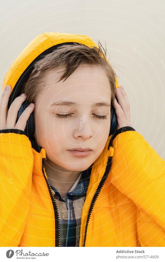 A contemplative young boy in a striking yellow jacket enjoys music on his headphones against a soft neutral background listening enjoyment peaceful serene