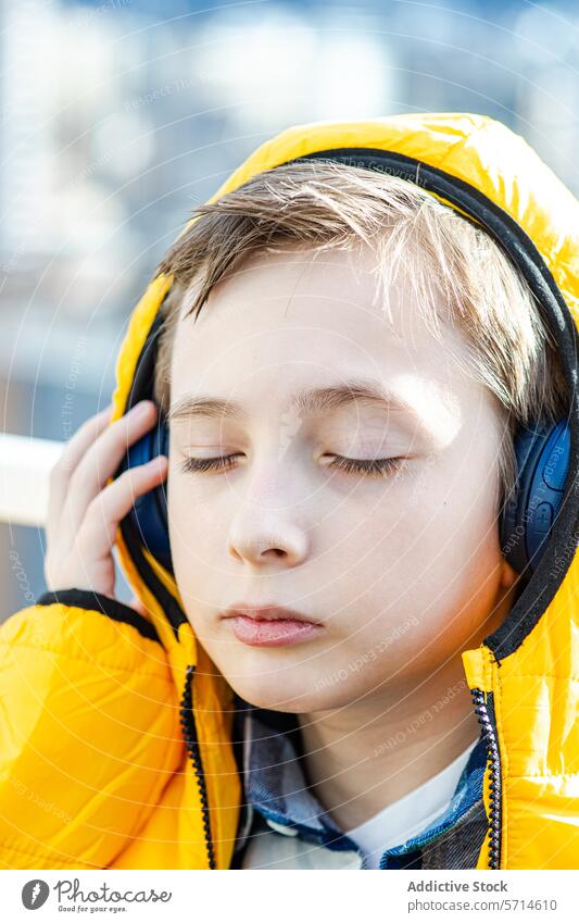 A serene boy with closed eyes enjoys music through black headphones, wearing a vibrant yellow hooded coat listening enjoyment audio leisure relaxation peaceful
