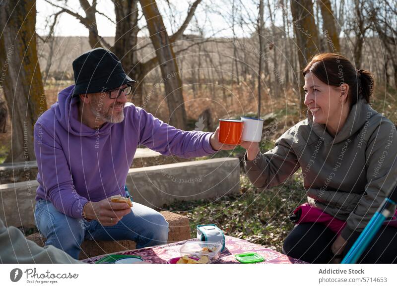 Two adults enjoying a picnic with a toast, smiling at each other while seated at a table with snacks, surrounded by trees smile family trek cup outdoors