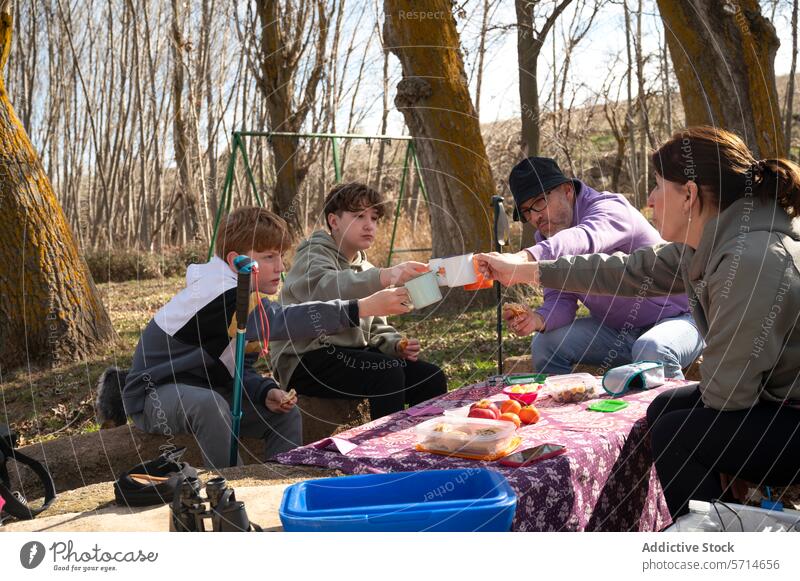 A family enjoys a picnic in the woods, toasting with cups as two children and two adults sit around a tablecloth-covered table with food outdoor gathering snack