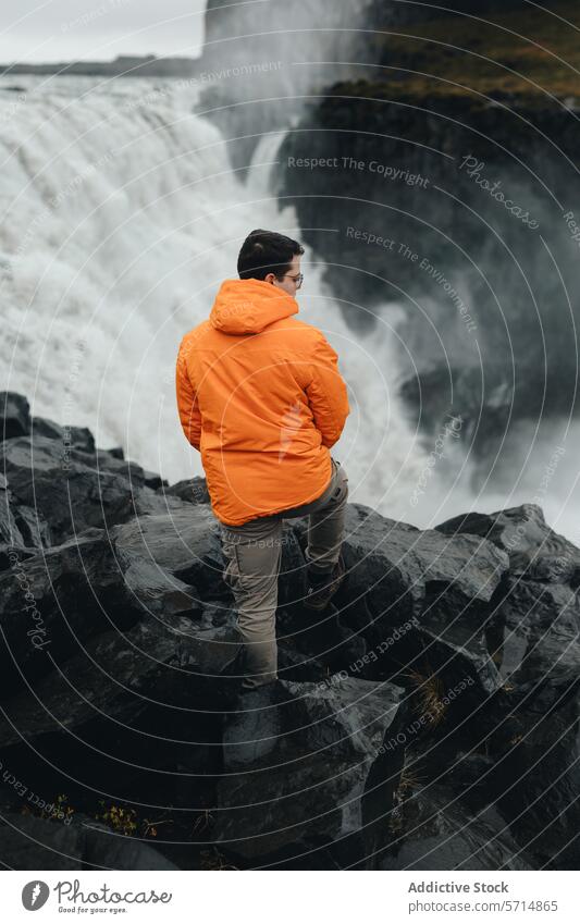 Anonymous man in vibrant orange jacket gazes at Gullfoss Waterfall, Iceland traveler male iceland gullfoss waterfall volcanic rocks nature outdoors adventure