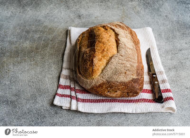 Top view of golden-brown rye sourdough bread on a white cloth with red stripes, next to a bread knife on a textured surface Sourdough homemade striped baking