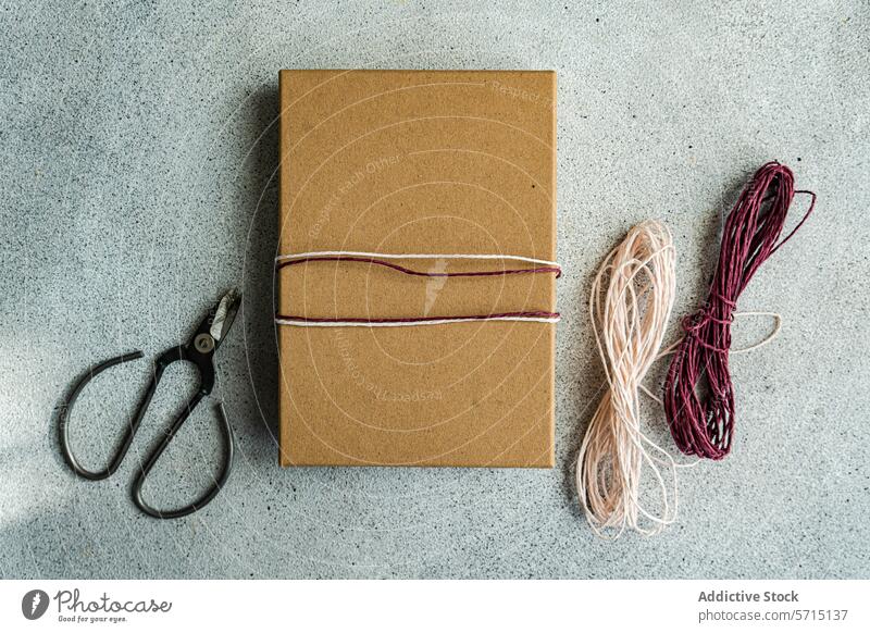 A simple yet elegant gift wrapped in brown paper with a pink and white twine, alongside scissors and additional twine on a textured grey background box wrapping