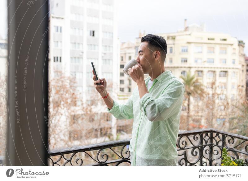 Asian man enjoying coffee while using smartphone on balcony asian urban outdoor daylight young adult male city leisure technology mobile casual lifestyle