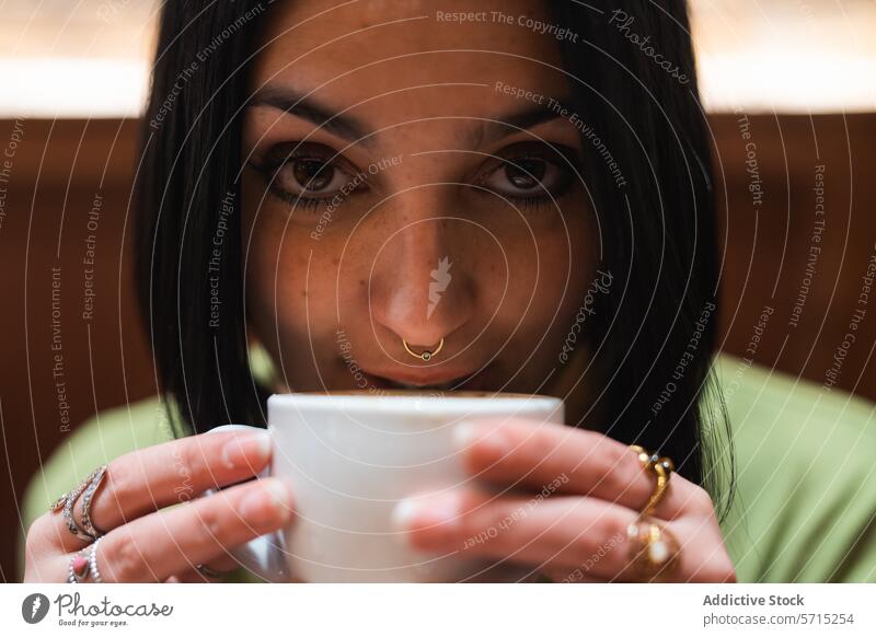 Intimate moment of woman enjoying her coffee cup close-up gaze intimate pensive jewelry nose ring warmth savor delicate young adult human face eyes focus hands