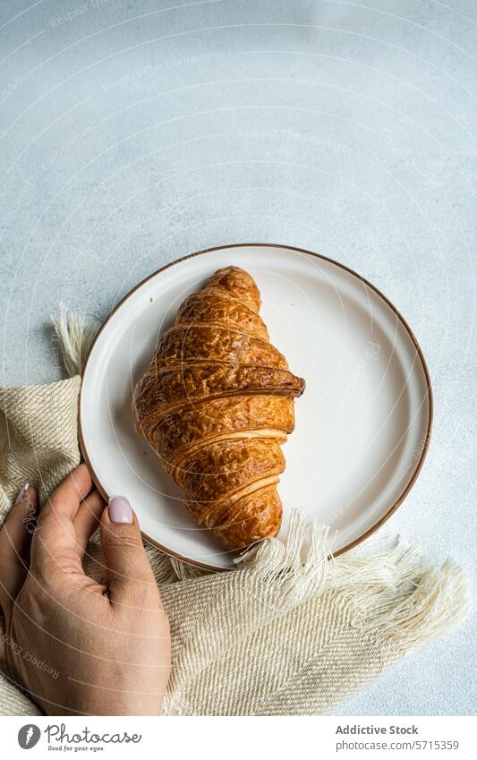 Top view of anonymous person hand holding a plate with a freshly baked croissant on a textured cloth, capturing a simple and cozy breakfast moment food morning
