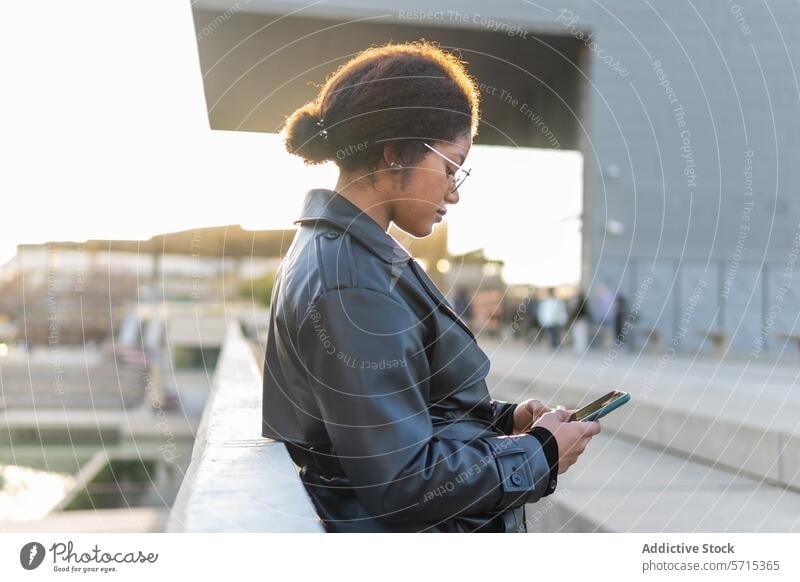 A woman wearing a winter coat and glasses is focused on her smartphone while standing on a city bridge during sunset Woman outdoor lifestyle urban technology