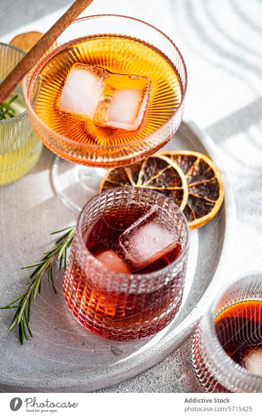 An array of exquisite alcoholic beverages in decorative glasses, accompanied by ice cubes, a cinnamon stick, rosemary, and dried citrus slices on a marbled tray