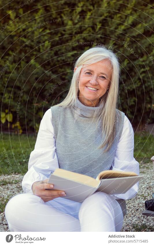 Smiling mature woman sitting outdoors reading a book, with a camera beside her, depicting an active and engaged lifestyle Woman smiling 50s leisure happy